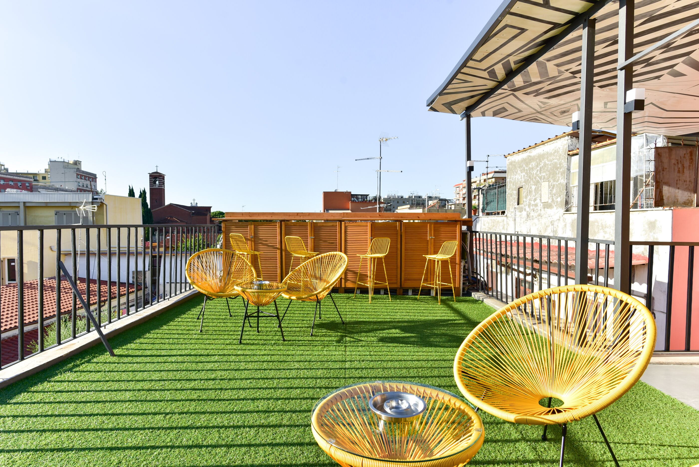 Una terrazza sul tetto con erba sintetica, sedie in vimini gialle e vista sulla città.