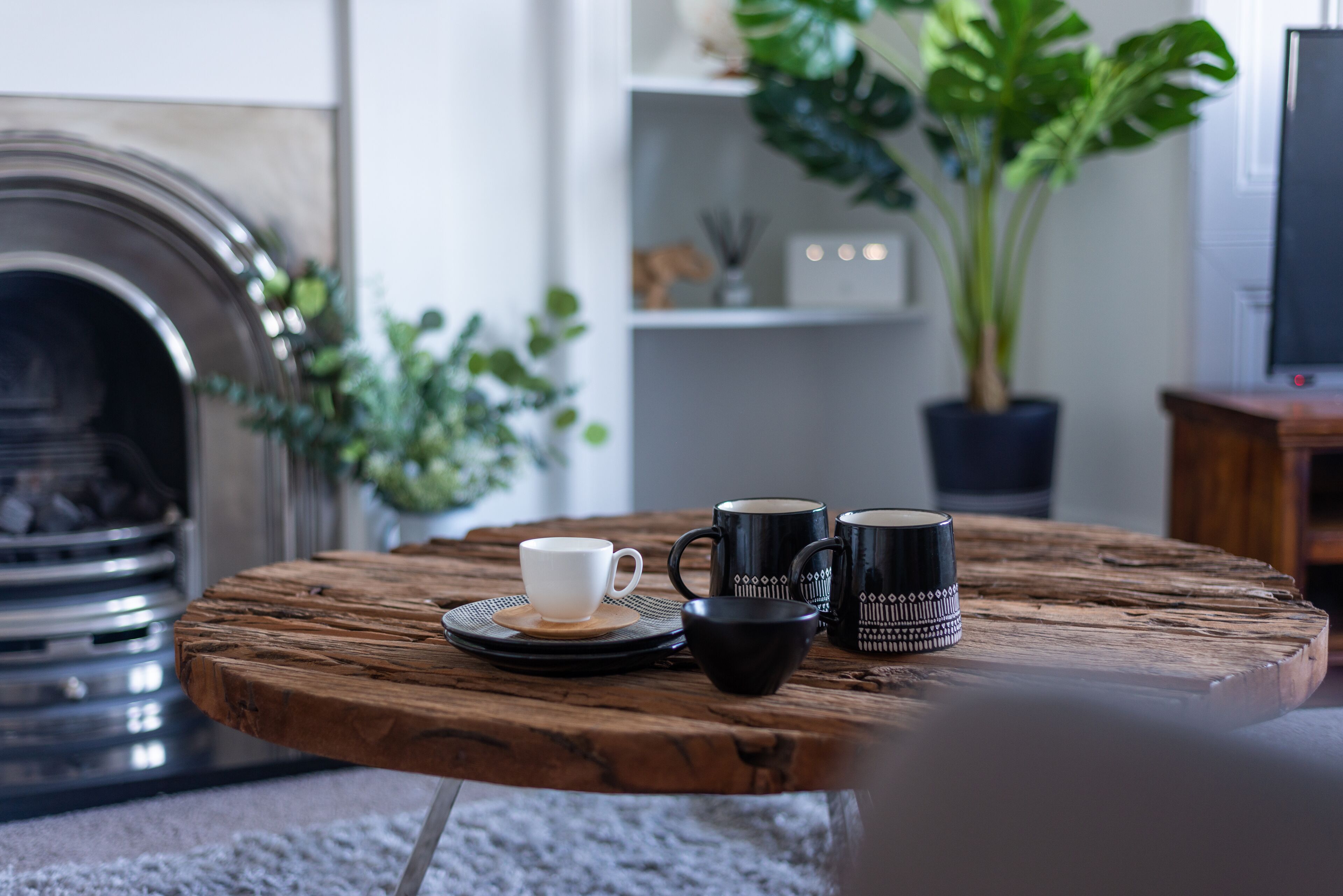 A round wooden table with a white cup, two black mugs, and a black cup on a coaster.