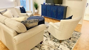 Una sala de estar con sofá, sillón, reposapiés y televisión sobre un mueble.