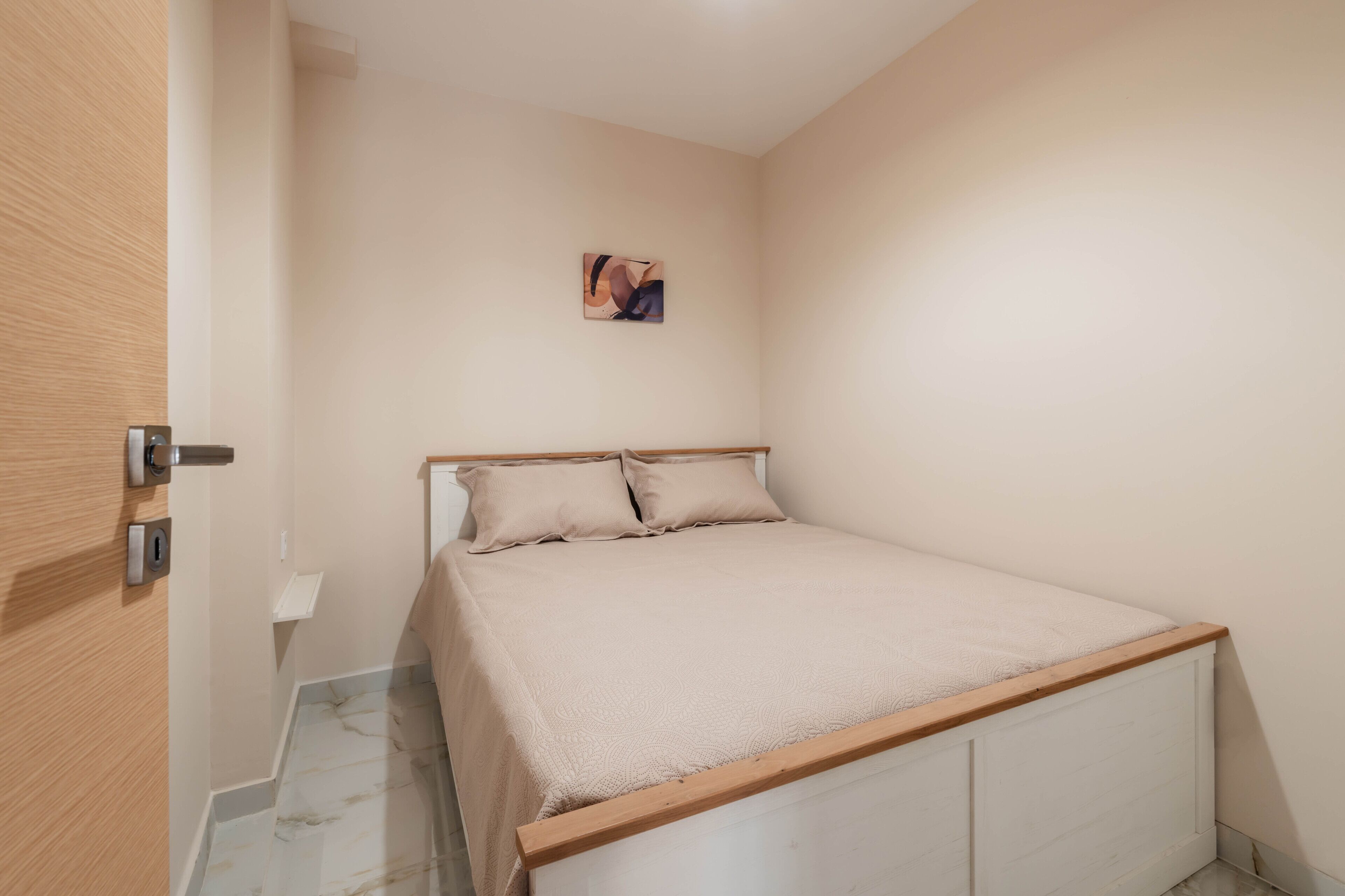 A small, neatly arranged bedroom with a single bed, a wooden door, and a small painting on the wall.