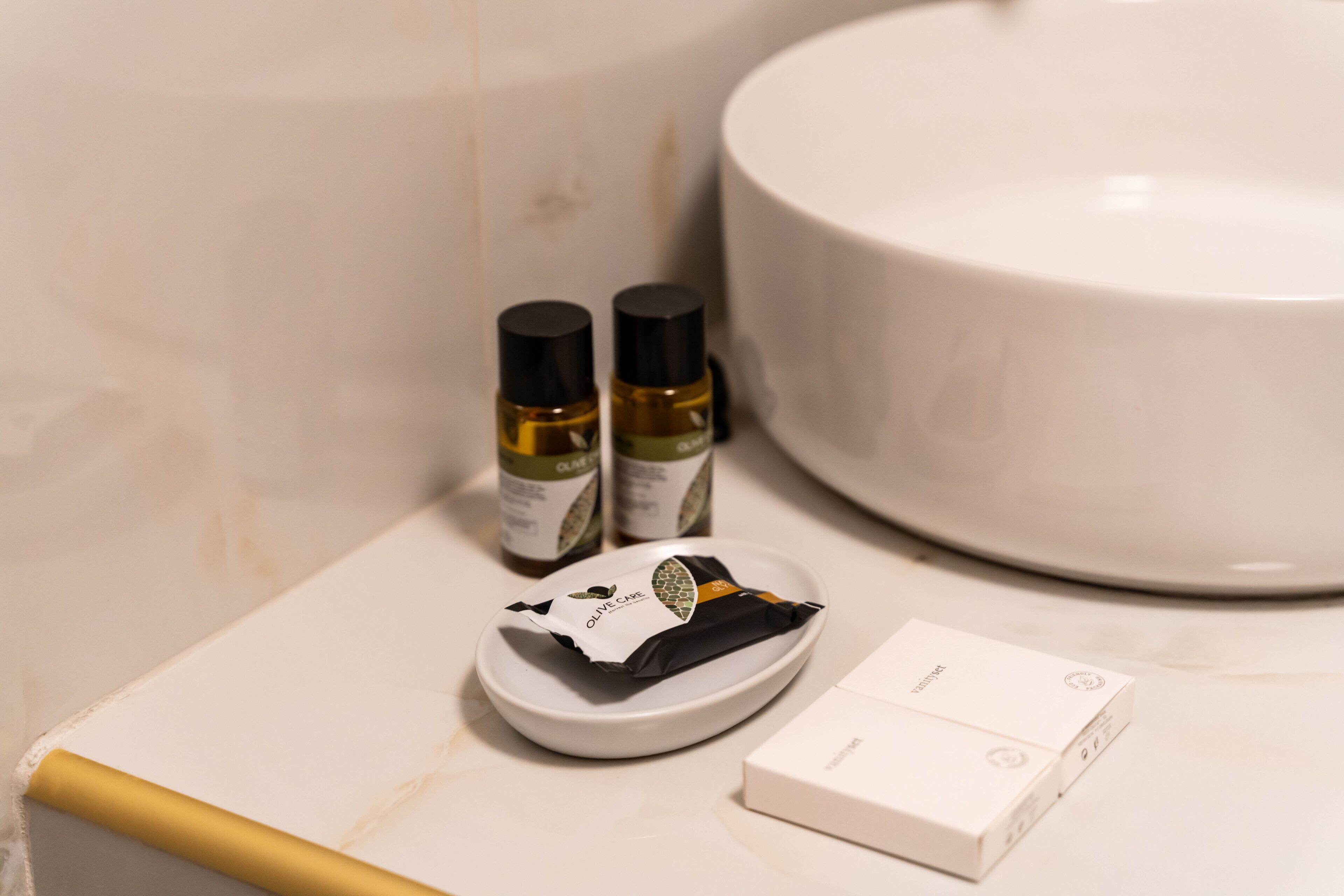 A bathroom with a white sink, two bottles of olive oil, a black and white toiletry pouch, and a white box on the counter.