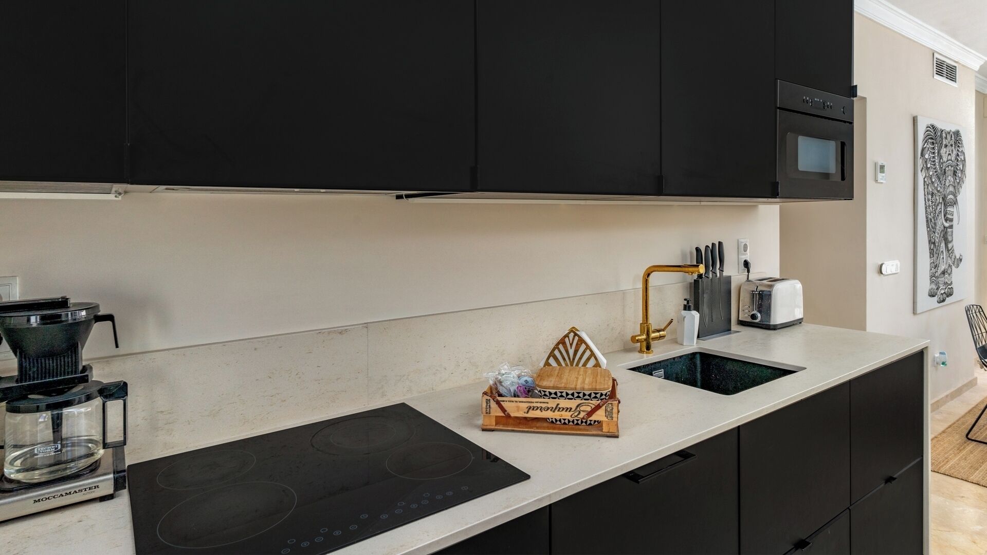 Een moderne keuken met zwarte kastjes, een ingebouwde oven, een koffiezetapparaat en een wastafel met een gouden kraan.