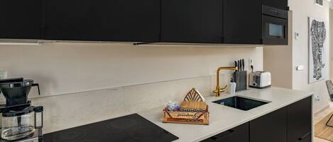 Een moderne keuken met zwarte kastjes, een ingebouwde oven, een koffiezetapparaat en een wastafel met een gouden kraan.