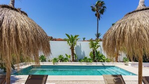 Een zwembad met rieten parasols, palmbomen en een witte muur op de achtergrond.