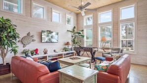 A spacious living room with a wooden ceiling, large windows, a ceiling fan, and a red leather sofa. - "Escape to Palm SunDaze, where the soothing ocean breeze melts your cares away, embracing you in the tranquility of coastal living. (Port Aransas)
