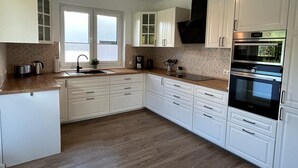 A modern kitchen with white cabinets, a wooden countertop, a built-in oven, and a window with blinds. - Exklusive Ferienwohnung bei Berlin, bis 5 Gäste, Große Terrasse, Garten (Bestensee)