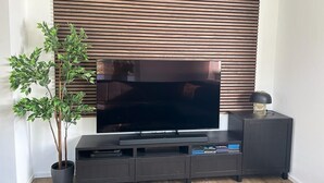 A modern living room with a flat-screen TV mounted on a wall, a dark entertainment unit below, and a potted plant to the left. - Exklusive Ferienwohnung bei Berlin, bis 5 Gäste, Große Terrasse, Garten (Bestensee)