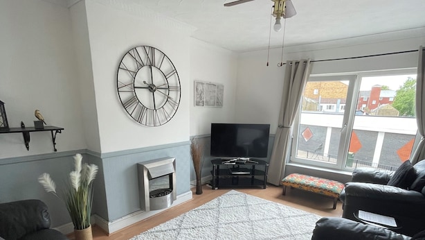 A living room with black leather sofas, a large clock, a flat-screen TV, and a ceiling fan. - March-2bed Apt - Family Friendly - Pet Friendly (March)