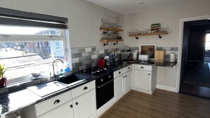 A modern kitchen with white cabinets, a black countertop, and a wooden floor. There are two wooden shelves above the counter holding glassware. - March-2bed Apt - Family Friendly - Pet Friendly (March)