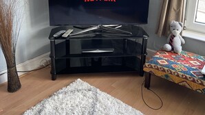 A living room with a large flat-screen TV on a black stand, a teddy bear on a small table, and a large fluffy rug on the floor. - March-2bed Apt - Family Friendly - Pet Friendly (March)