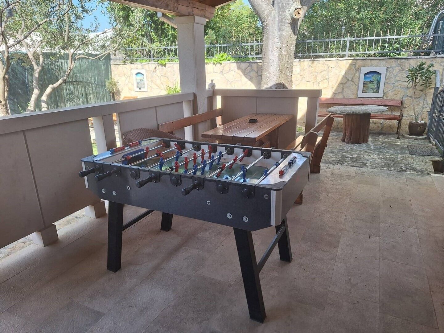 Een terras buiten met een tafelvoetbal tafel, houten banken en een stenen muur op de achtergrond.