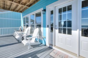 A covered porch with white Adirondack chairs, a blue exterior, and a white door with a window. - Enjoy ocean views at The Hawks Nest, a spacious 5-bedroom home in Oak Island. (Oak Island)