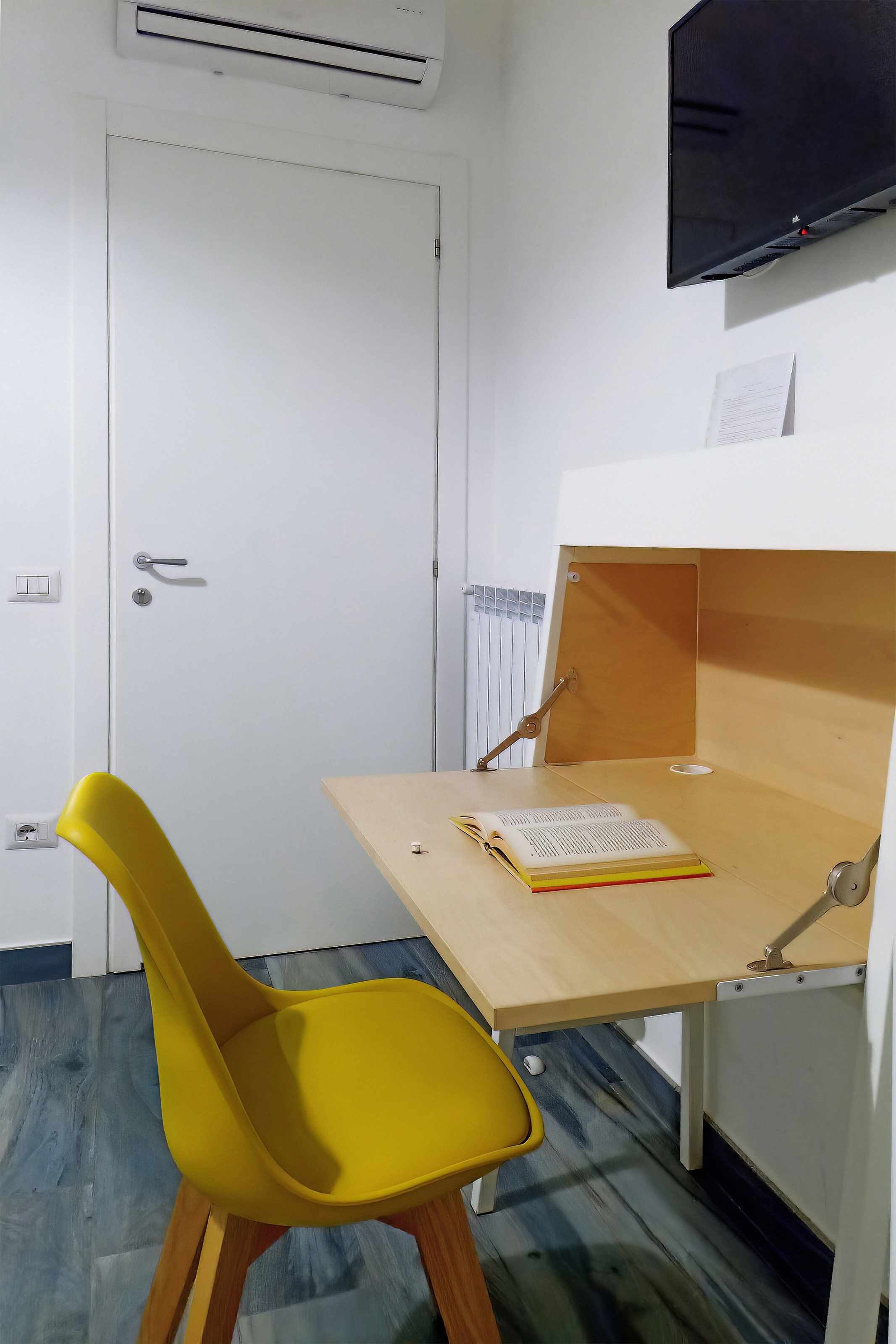 A small room with a wooden desk, a yellow chair, a closed door, and a mounted TV.