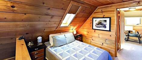 A wooden cabin bedroom with a bed, dresser, and a small table with a lamp.