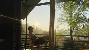 Una habitación con un amplio ventanal que da a un balcón con vistas a árboles y al atardecer.