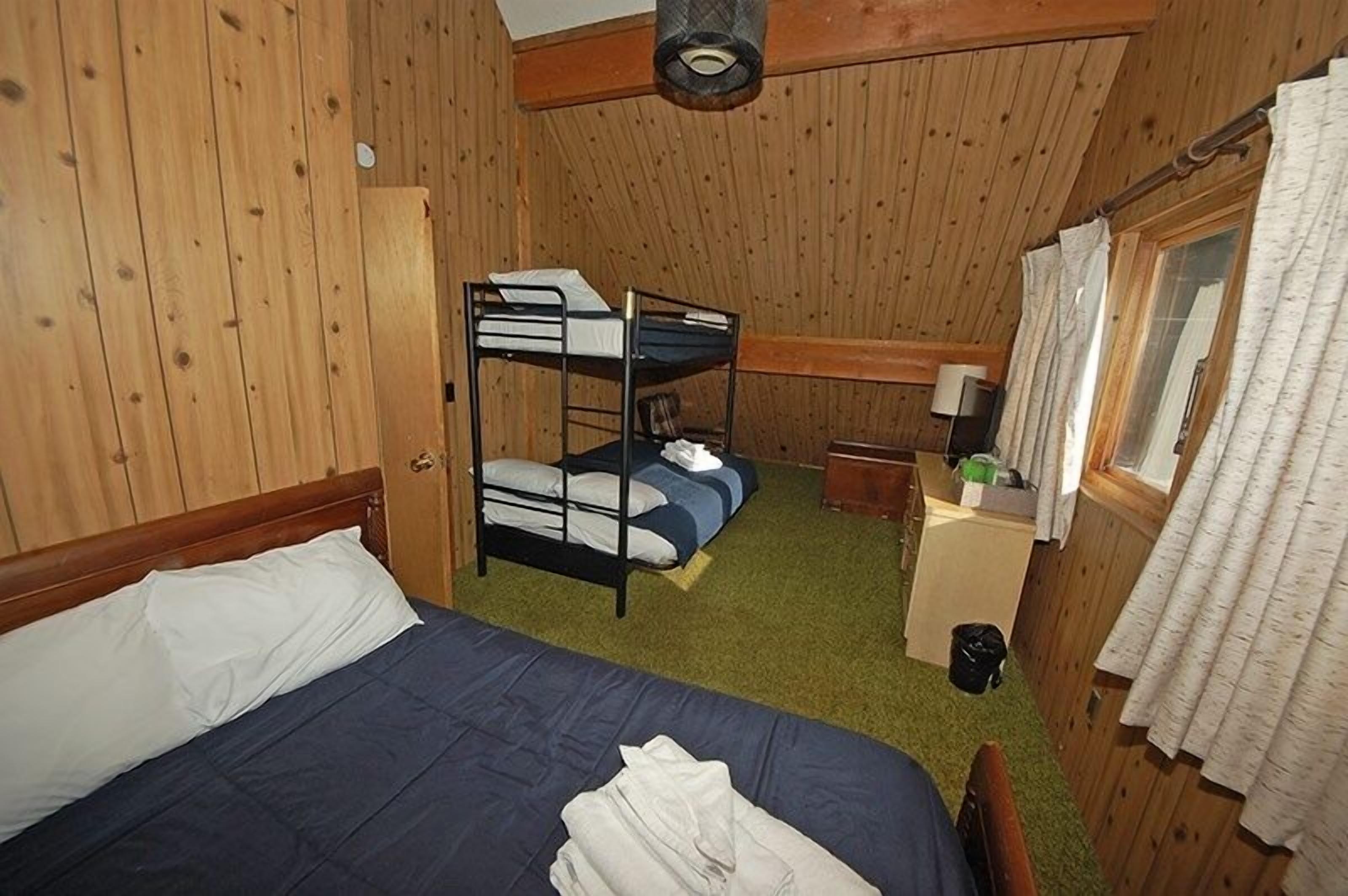 A bunk bed room with a wooden ceiling and walls, a single bed with a blue mattress, a small table with a lamp, and a window with curtains.