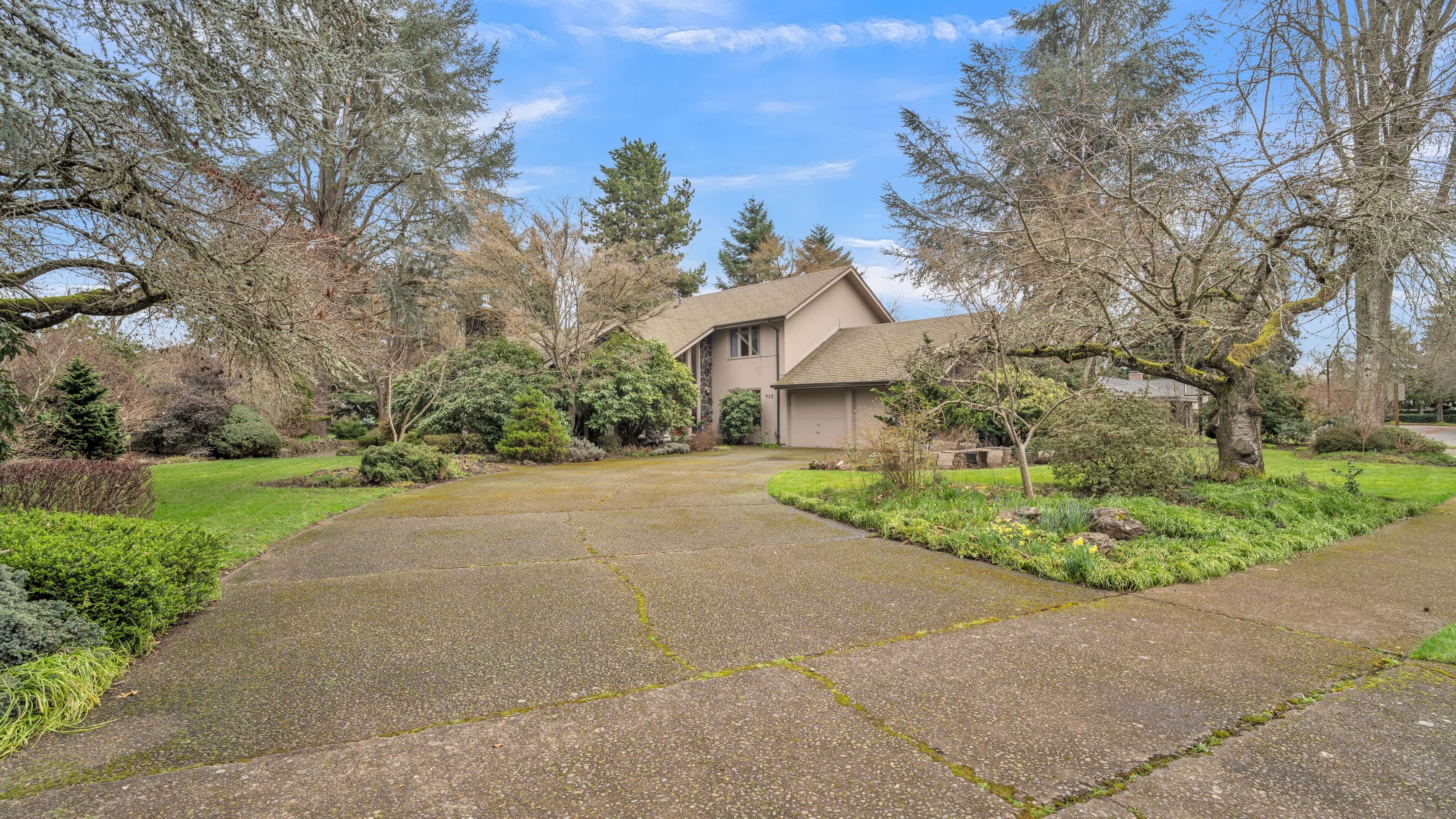A two-story house with a stone facade, a front yard with a driveway, and a variety of trees and shrubs.