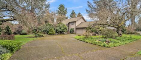 A two-story house with a stone facade, a front yard with a driveway, and a variety of trees and shrubs.