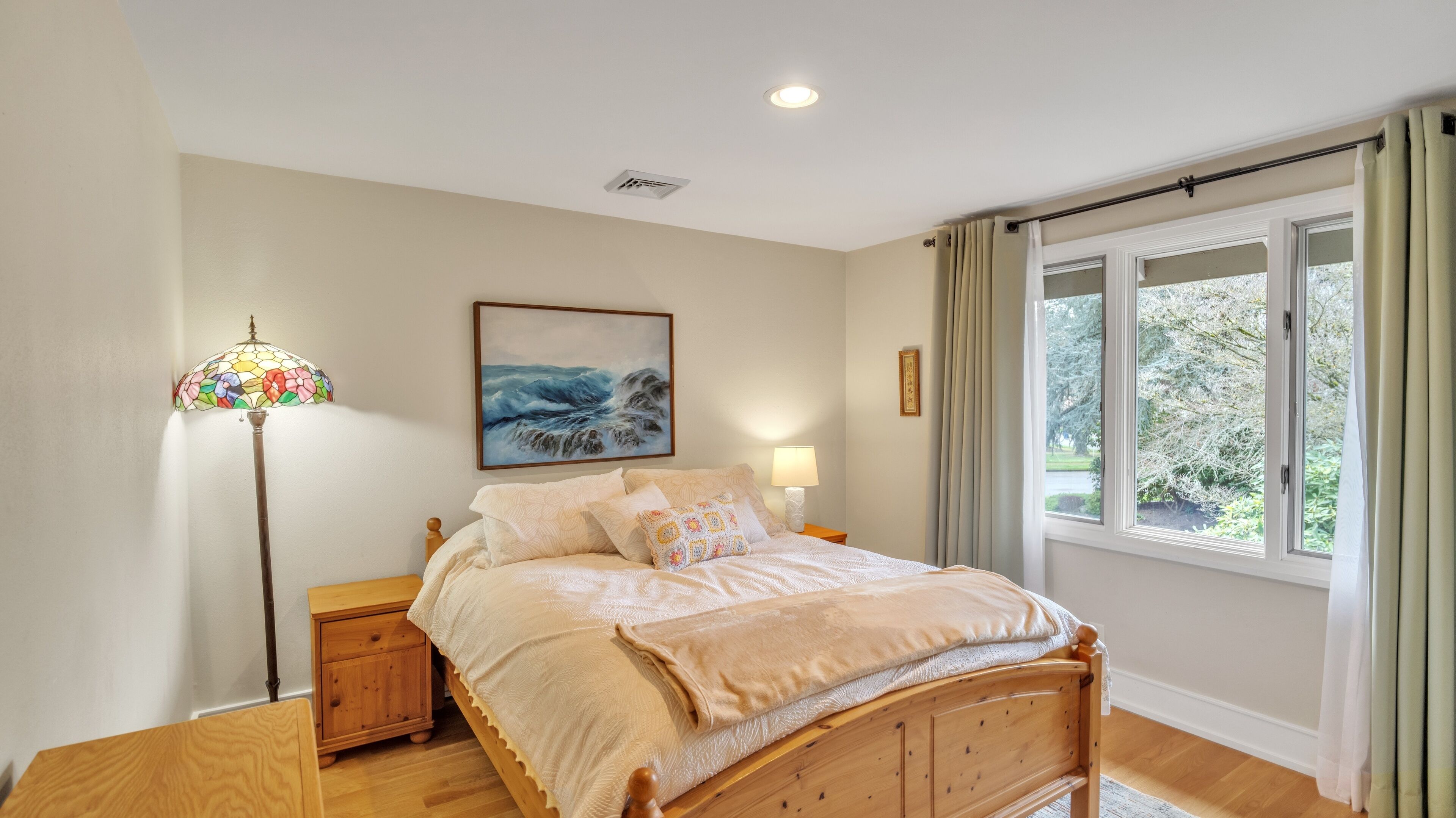 A bedroom with a wooden bed, a nightstand, a wall-mounted lamp, a painting, and a window with curtains.