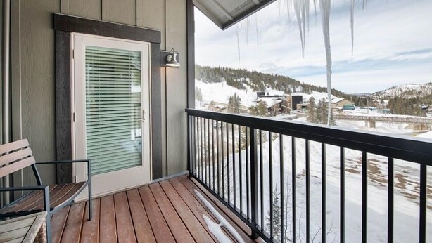 A wooden deck with a bench and a door with blinds, overlooking a snowy landscape with buildings and a bridge. - Select Unit 3606 - Fraser Crossing/founders Pointe 2 Bedroom Condo (Winter Park)