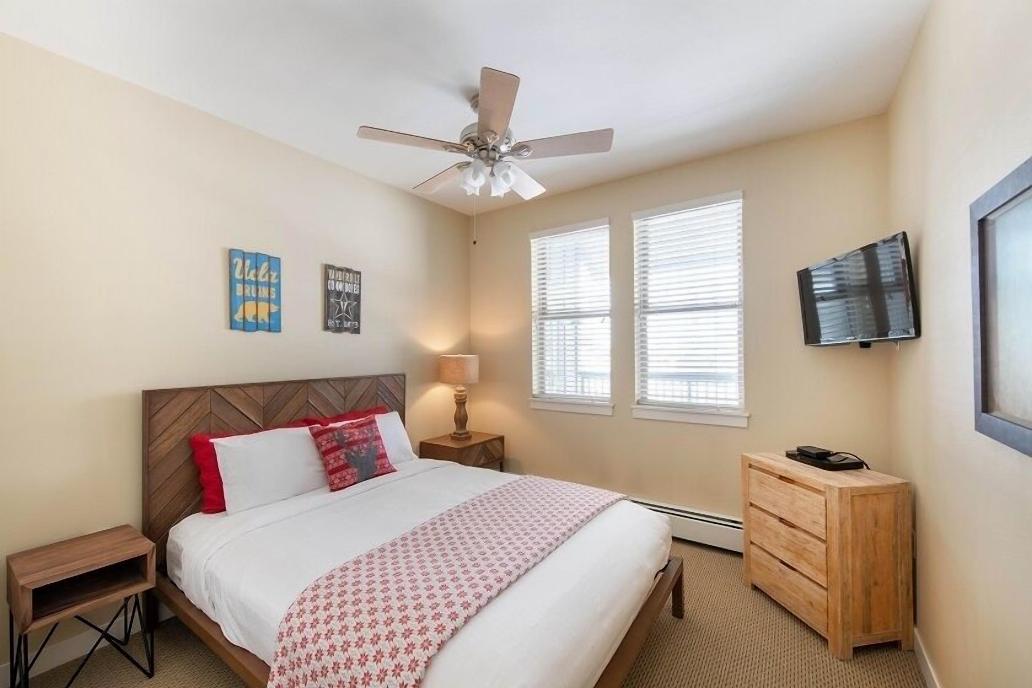 A bedroom with a bed, a nightstand, a dresser, a ceiling fan, and a TV mounted on the wall.
