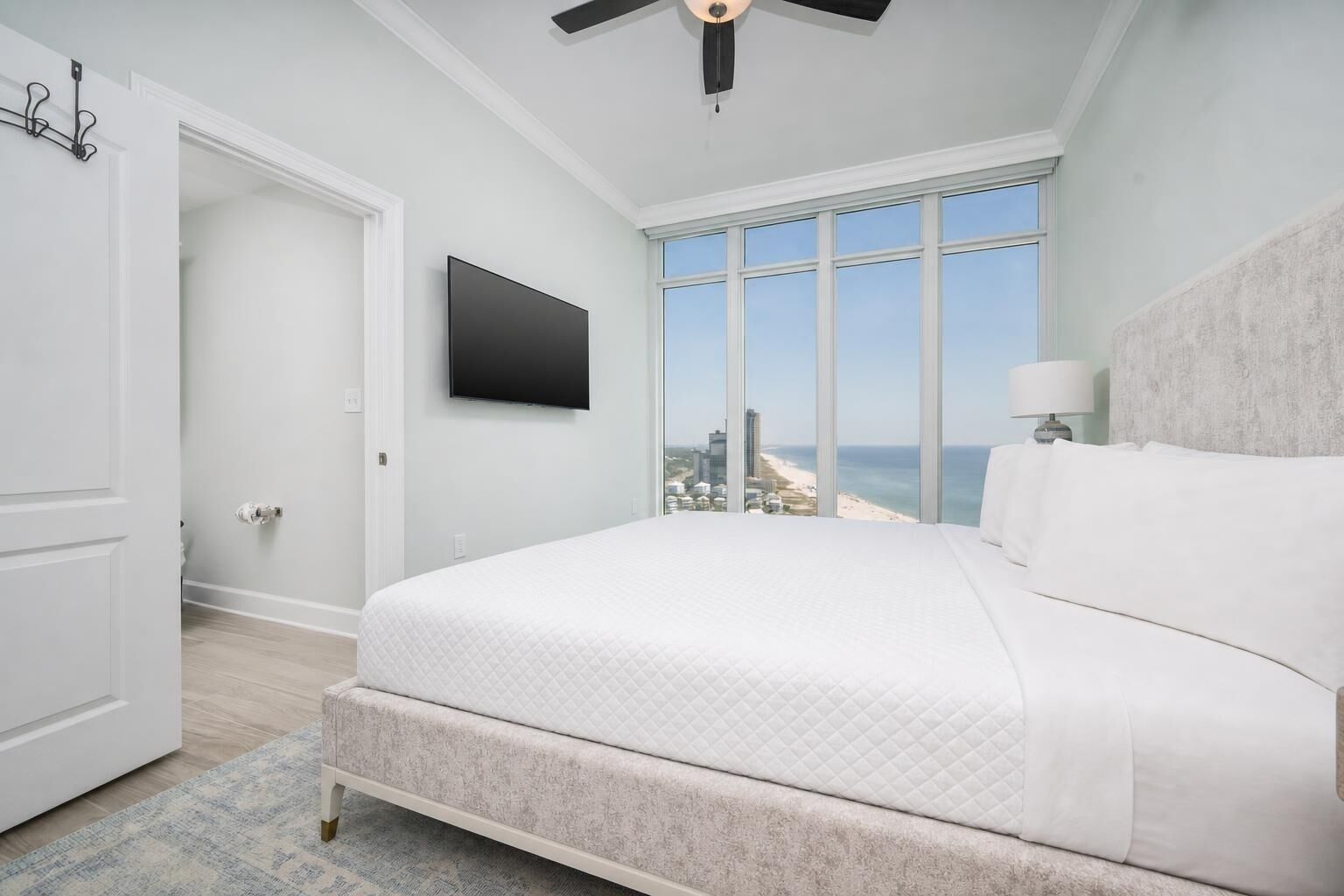 A bedroom with a large bed, a TV, a ceiling fan, and a view of the beach.