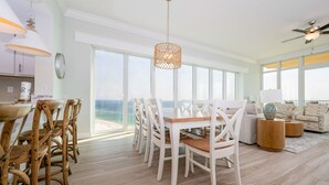 A spacious dining area with a view of the ocean, a cozy seating area with a sofa and side tables, and a kitchen in the background.