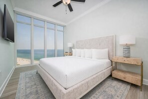 A bedroom with a large bed, a ceiling fan, a TV, a nightstand, and a view of the beach. - Phoenix Gulf Towers 2108 4 Bedroom Condo (Orange Beach)