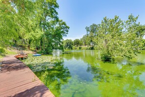 A wooden deck leading to a calm pond surrounded by lush greenery and trees. - Seguin Riverfront Retreat: Fishing Dock + Kayaks! (Seguin)