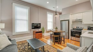 A modern living room with a gray sofa, a coffee table, and a patterned rug. There is a dining area with a wooden table and chairs. The kitchen features white cabinets, a stainless steel refrigerator, and an oven. - Downtown Hot Springs Condo - Walk to Restaurants! (Hot Springs)