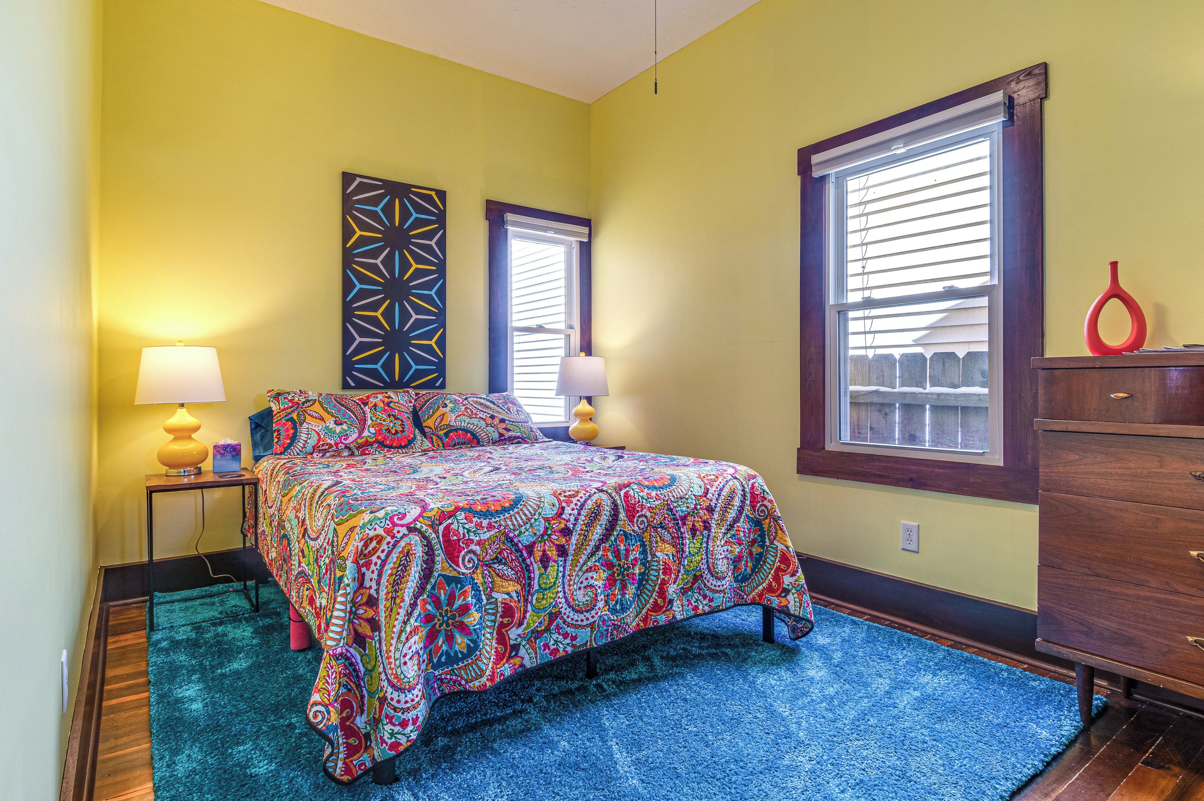 A bedroom with a colorful bedspread, two bedside lamps, a dresser, and a window with blinds.