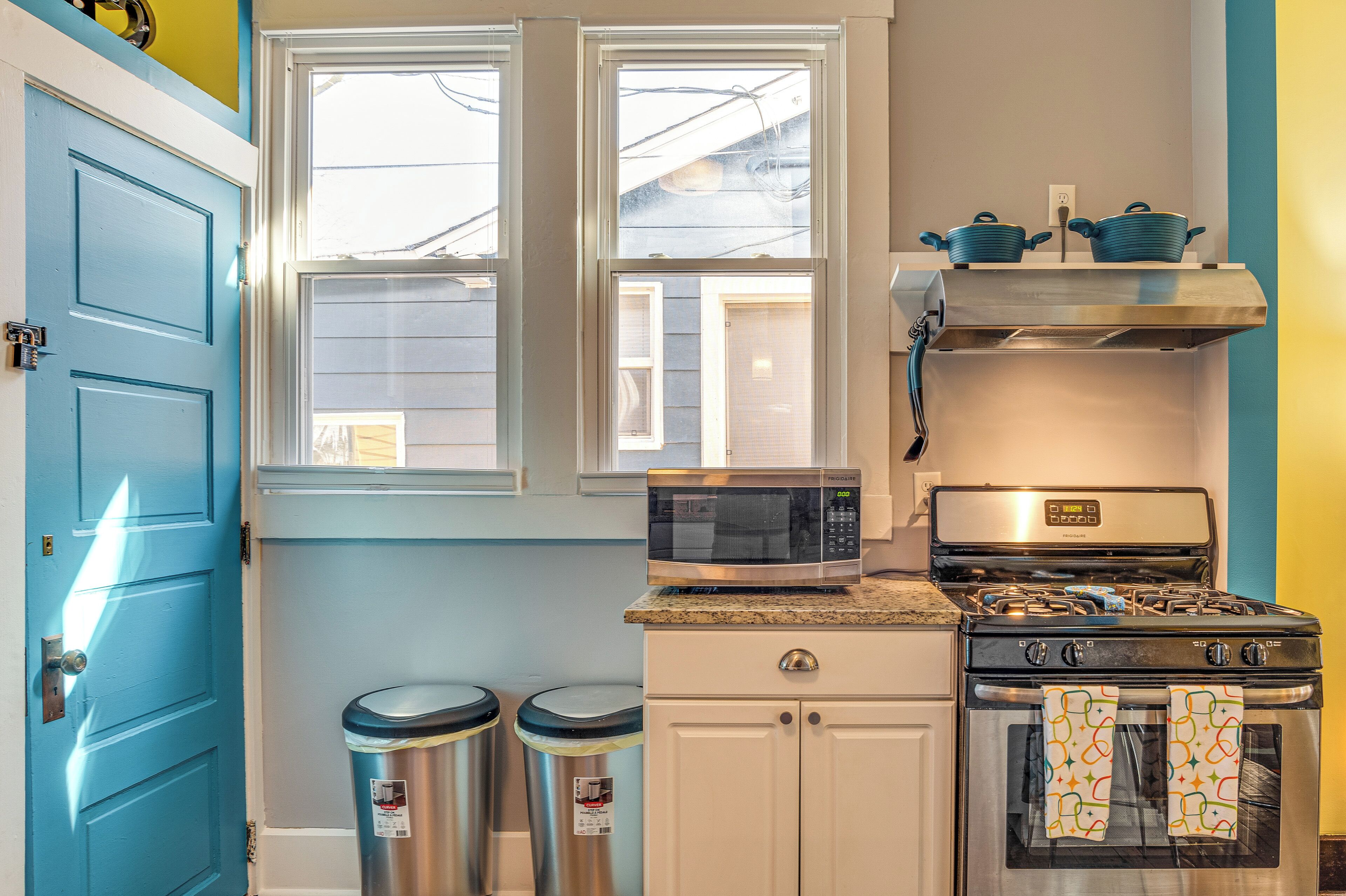 A kitchen with a blue door, two windows, a microwave, a stove, and two trash cans.