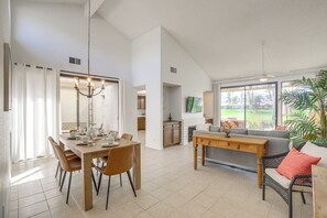 A spacious living area with a dining table, chairs, a sofa, and a large window offering a view of the outdoors. - Palm Desert Condo w/ Patio & Golf Course Views (Palm Desert)