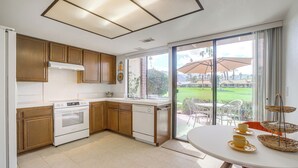 A modern kitchen with wooden cabinets, a white stove, and a dishwasher. There is a dining area with a white table and yellow cups. - Palm Desert Condo w/ Patio & Golf Course Views (Palm Desert)