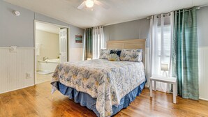 Ein Schlafzimmer mit Bett, Deckenventilator, Nachttisch und einem durchgehenden Türrahmen einsehbaren Badezimmer.
