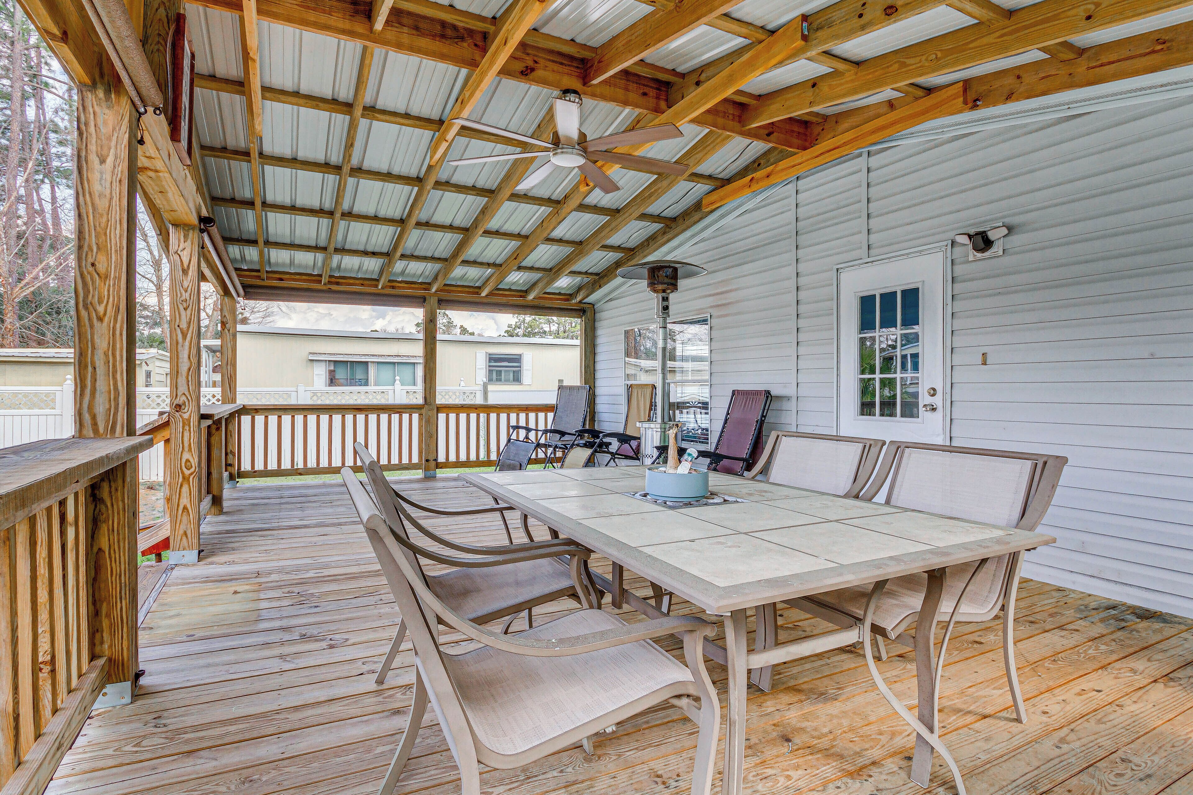 A covered outdoor patio with a wooden table and chairs, a ceiling fan, and a view of a house in the background.