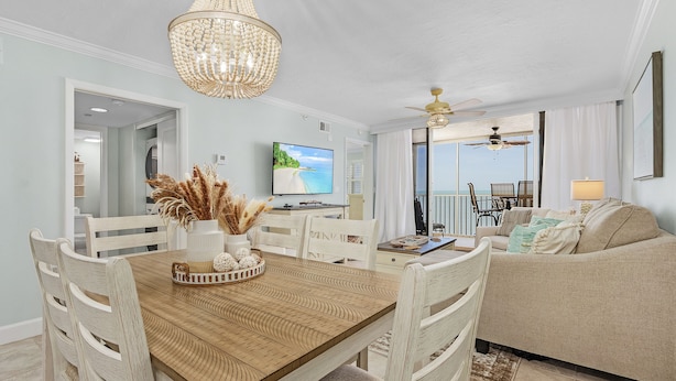 A dining area with a wooden table, white chairs, and a chandelier. There is a television on the wall and a view of the beach through the window. - Beach Villas # 505 - Bogo24 2 Bedroom Condo by RedAwning (Fort Myers Beach)