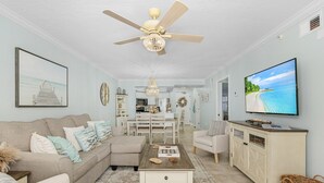 A living room with a sofa, coffee table, TV, and a ceiling fan. - Beach Villas # 505 - Bogo24 2 Bedroom Condo by RedAwning (Fort Myers Beach)