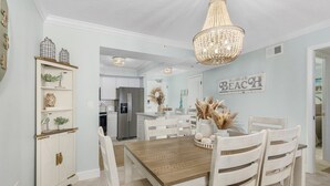 A dining area with a wooden table and white chairs, a chandelier, and a wall with a 'BEACH' sign. - Beach Villas # 505 - Bogo24 2 Bedroom Condo by RedAwning (Fort Myers Beach)