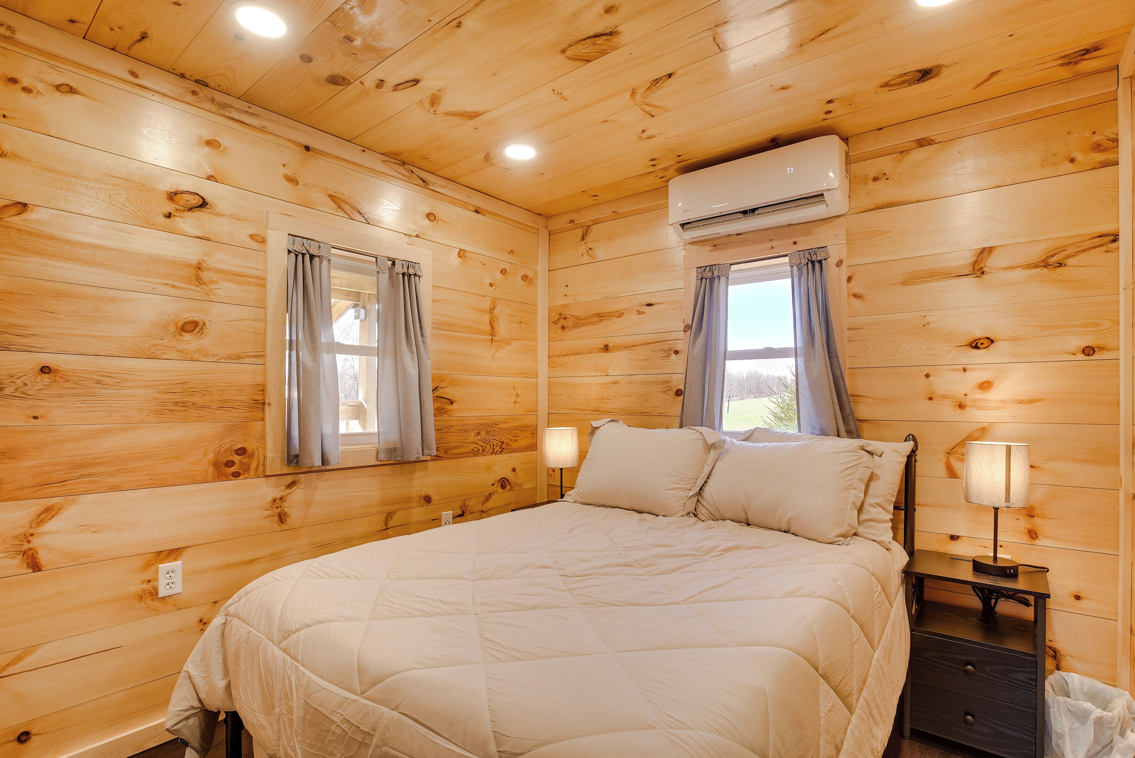 A wooden-paneled bedroom with a bed, two windows with curtains, an air conditioning unit, and a bedside table with a lamp.