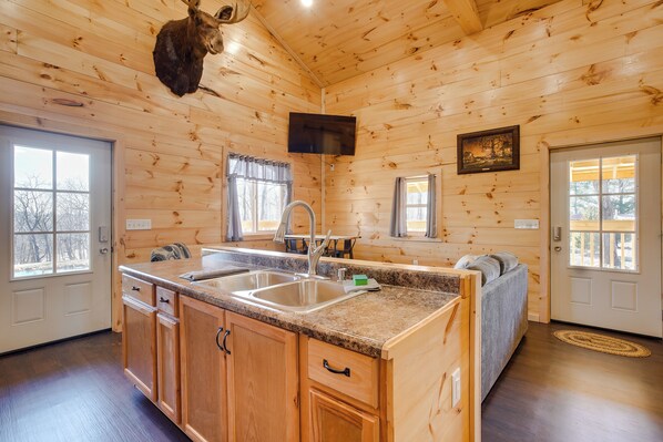 Interior de una cabaña de madera con cocina que incluye encimeras de granito, armarios de madera, un lavamanos y un televisor montado.
