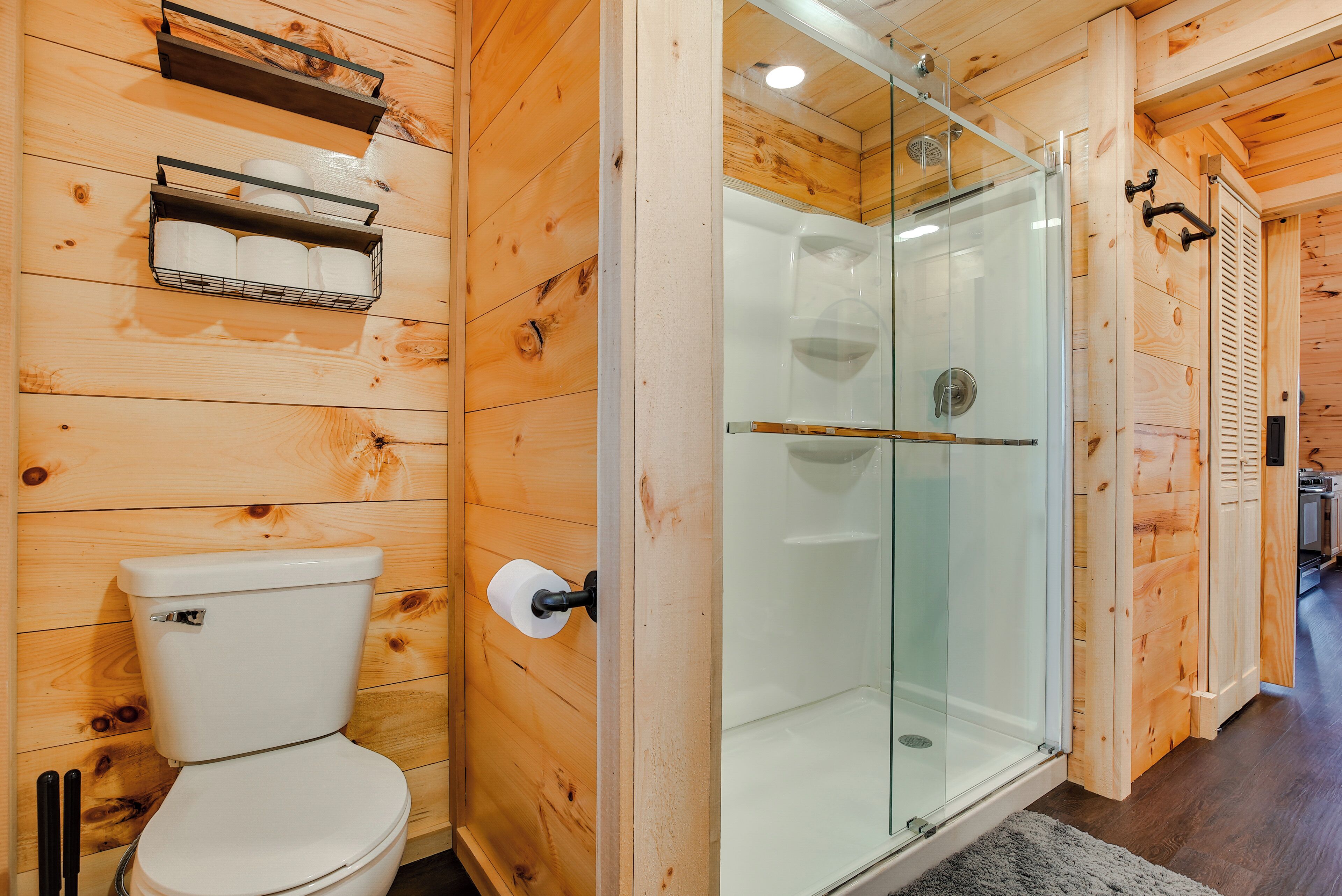 Un baño de madera con inodoro, ducha envuelta en vidrio y repisa con papel higiénico.