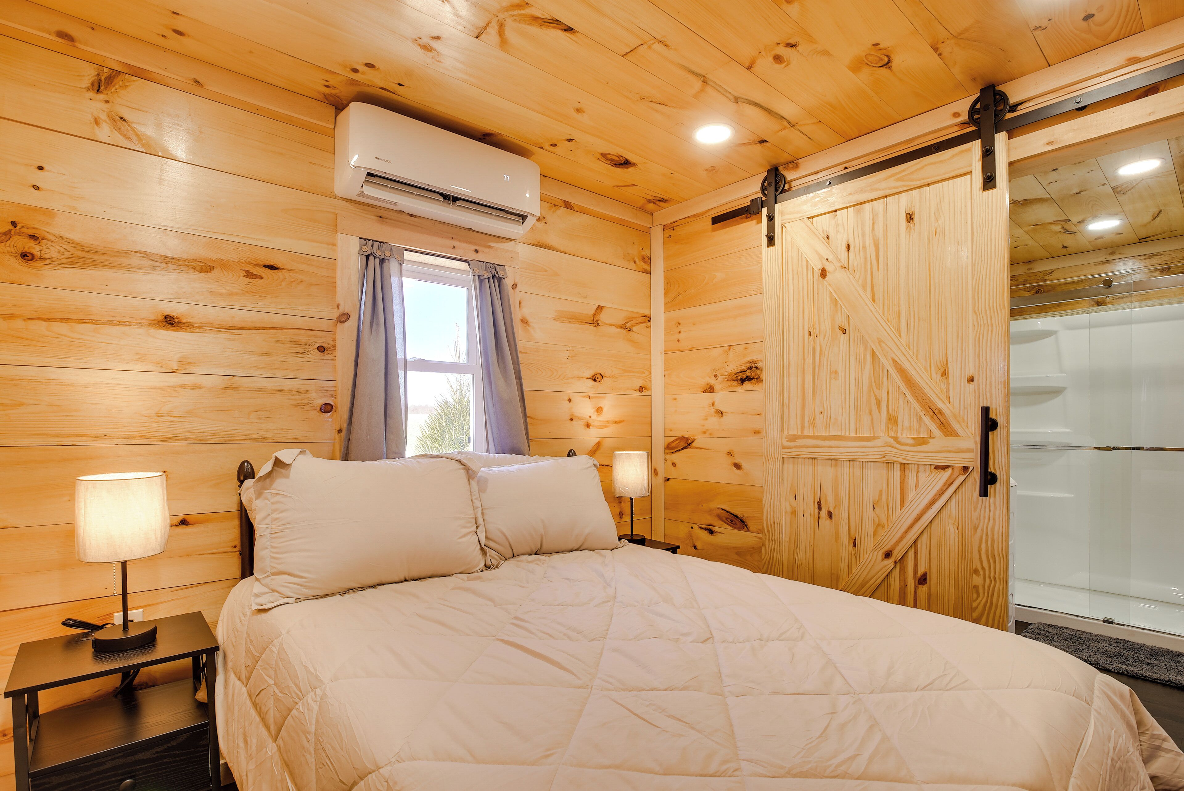 A wooden-paneled room with a bed, two bedside lamps, an air conditioning unit, and a sliding door to a bathroom.