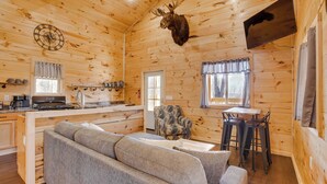 A wooden cabin interior with a sofa, dining table, chairs, a clock, a mounted moose head, and a kitchen area. - 'friends & Family' Rental Cabin w/ Hot Tub! (Somerset)