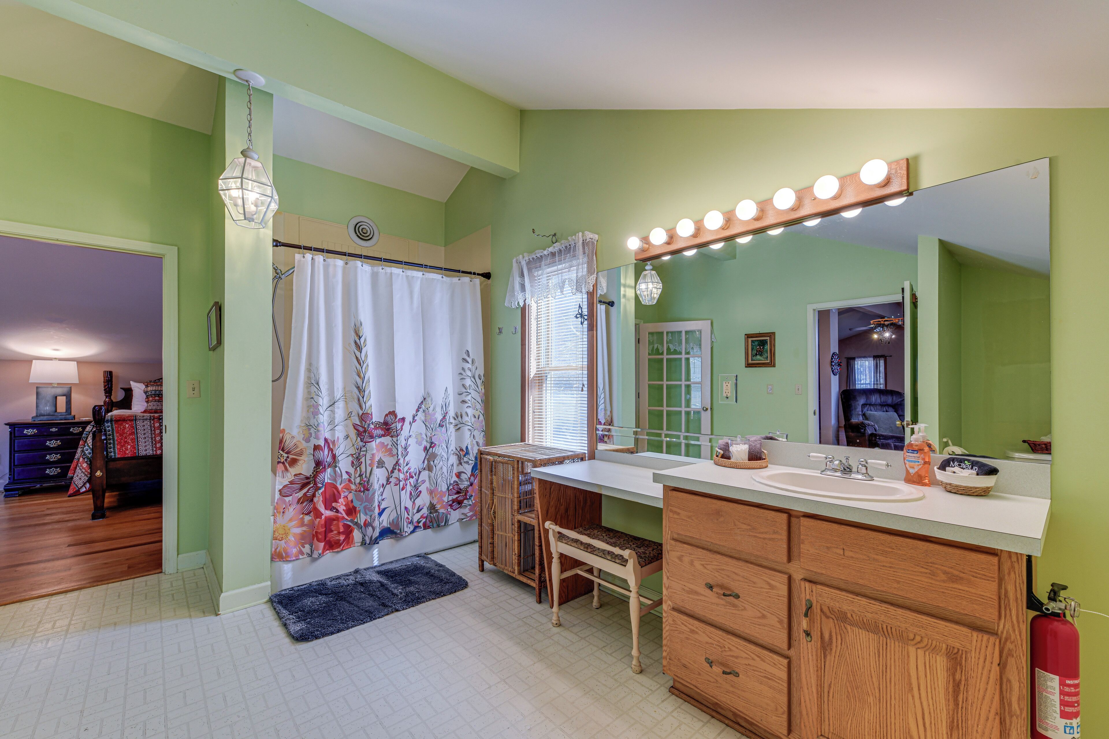 A bathroom with a wooden vanity, a large mirror with lights, a shower curtain, and a visible fire extinguisher.