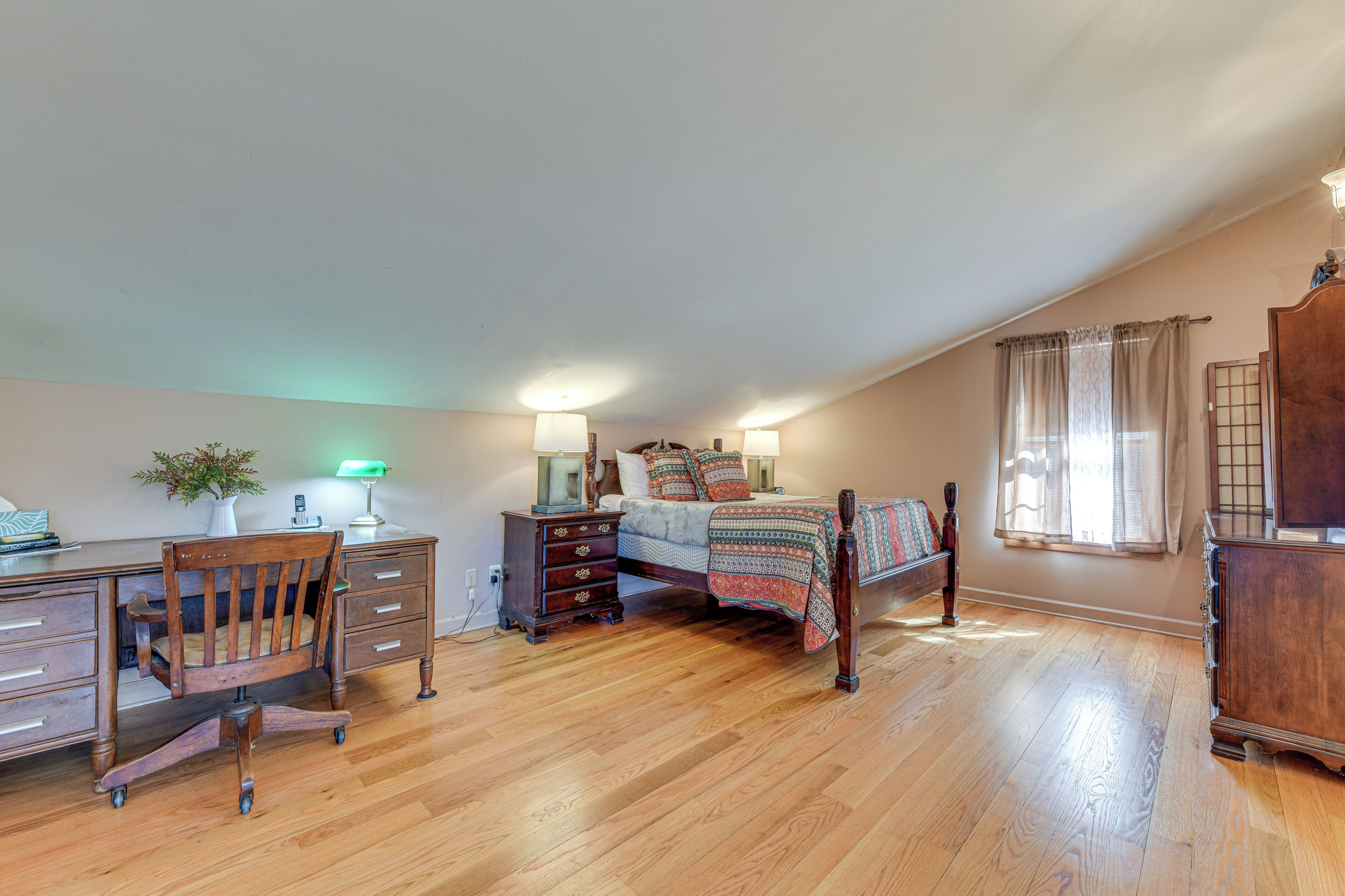 A bedroom with a wooden desk, a chair, a bed with a patterned blanket, a dresser, and a nightstand.
