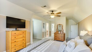 Quarto com cama, cómoda de madeira, televisão, ventoinha no teto e corredor que dá para outro espaço.