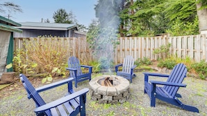 A fire pit with four Adirondack chairs around it, set on a gravel surface with a wooden fence and greenery in the background. - Rockaway Beach Home, Walk to Ocean, Pet Friendly! (Rockaway Beach)