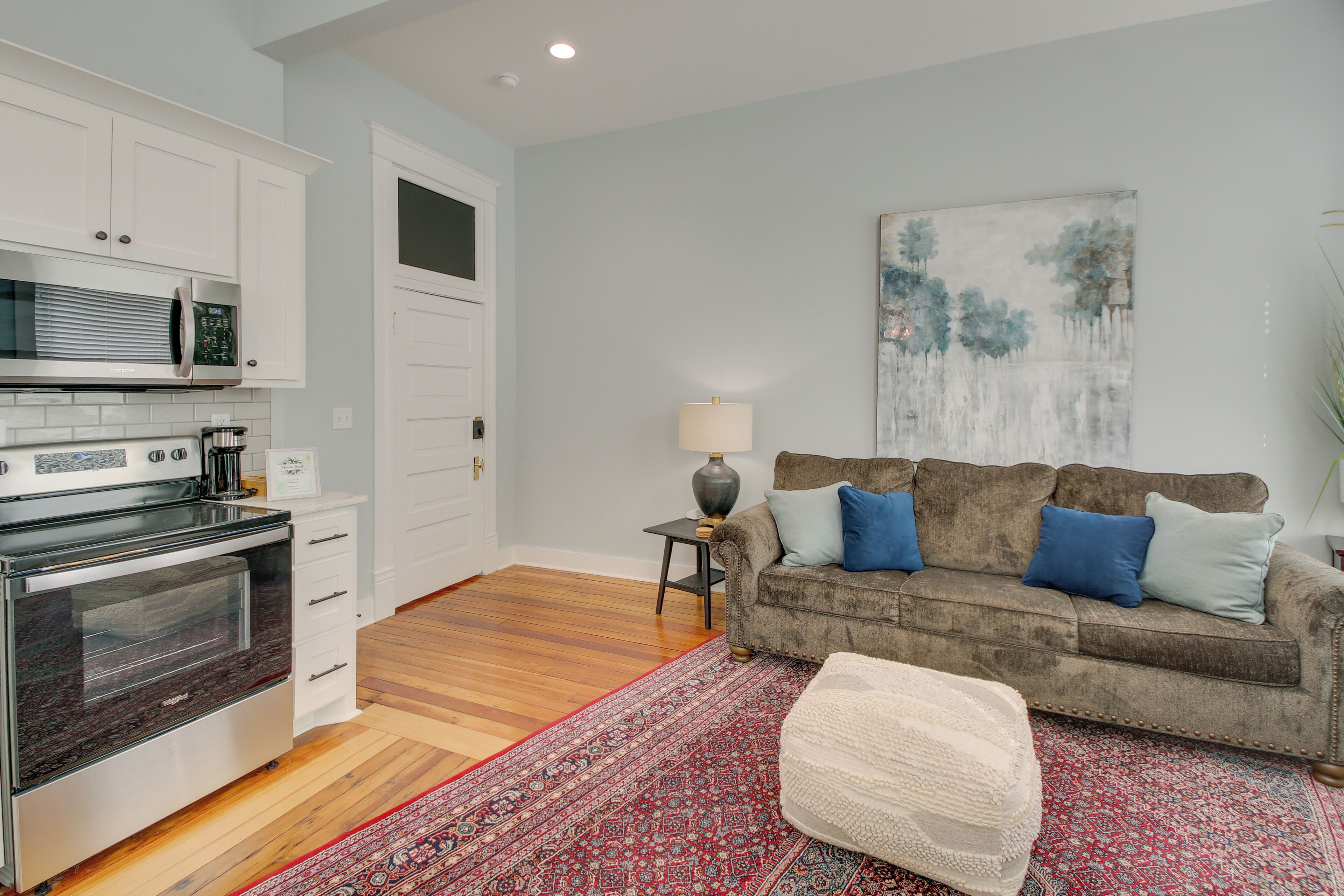 A modern kitchen with stainless steel appliances, a wooden floor, a red patterned rug, a beige ottoman, and a grey sofa with blue cushions.