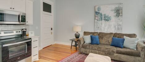 A modern kitchen with stainless steel appliances, a wooden floor, a red patterned rug, a beige ottoman, and a grey sofa with blue cushions.